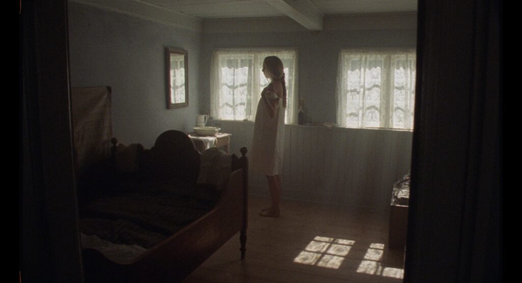 Young woman standing in a softly lit bedroom with two lace-curtained windows and a wooden bed.