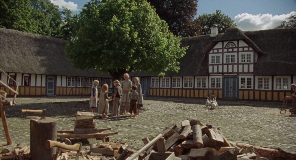 People gathered beneath a large tree in the courtyard of a historic Danish farmhouse, with timber and firewood in the foreground.