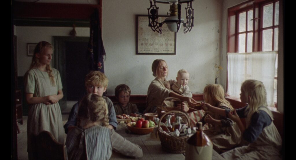 Family gathered around a dining table in a sunlit farmhouse interior with wooden windows and period furnishings.
