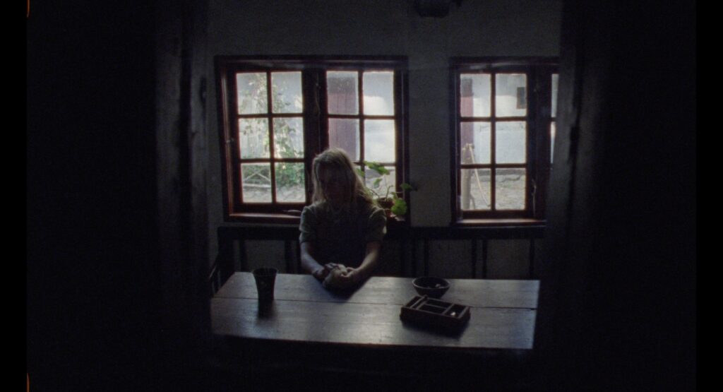 Young girl sitting alone at a wooden table facing two windows in a dark farmhouse interior.