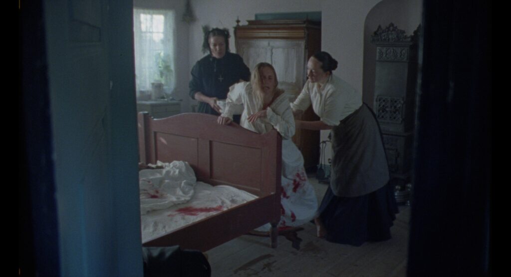 Women attending to a woman in bed inside a modest farmhouse bedroom with pale walls and wooden furniture.