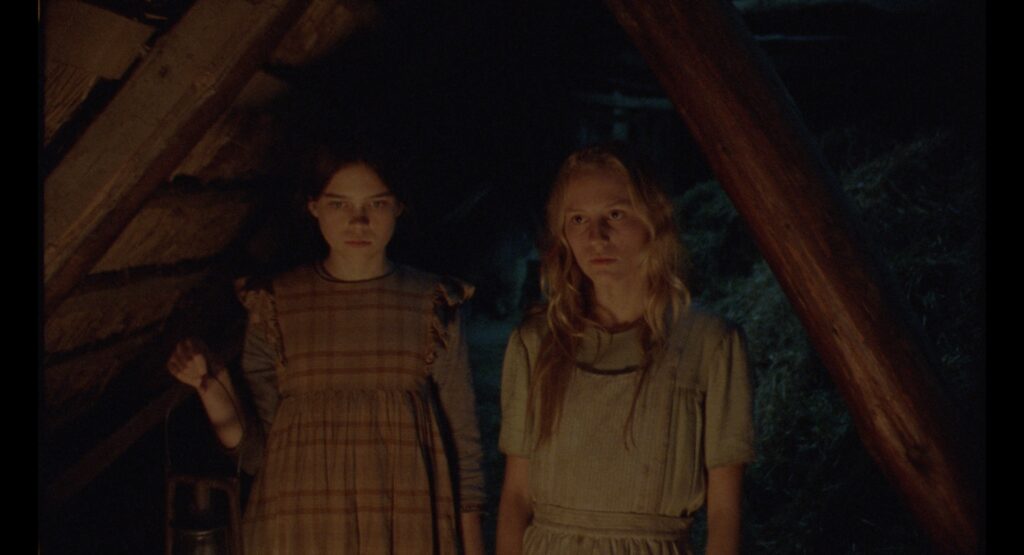 Two girls standing beneath exposed timber beams in a dark farmhouse attic.