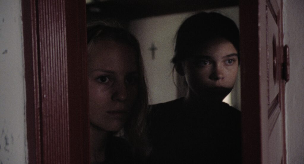Two young girls peering through a narrow doorway into a dimly lit farmhouse interior.