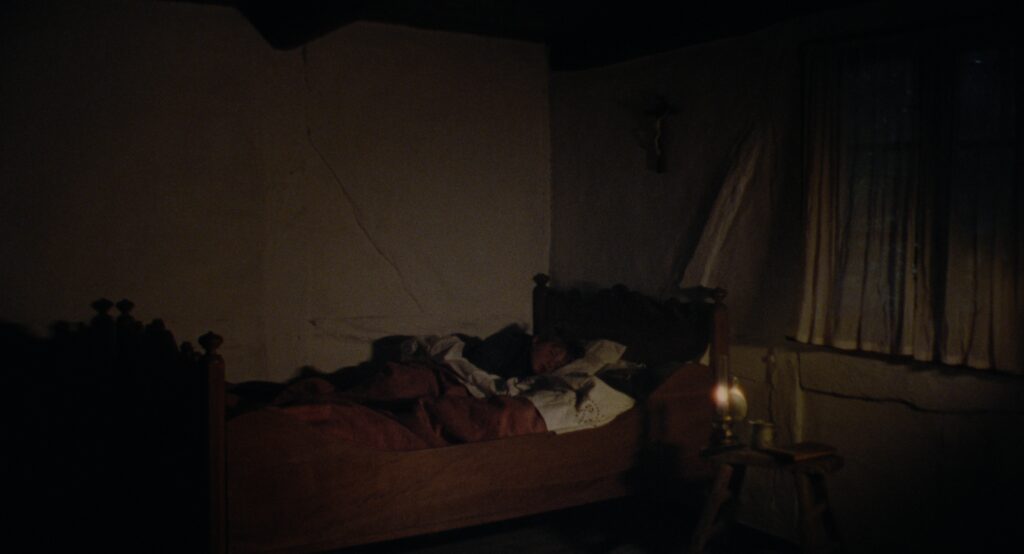 Dark farmhouse bedroom with a wooden bed, rumpled bedding and a small oil lamp casting warm light.