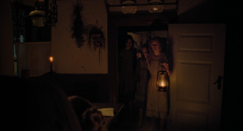 Young girls standing in a doorway holding an oil lantern, surrounded by the darkness of a farmhouse interior.