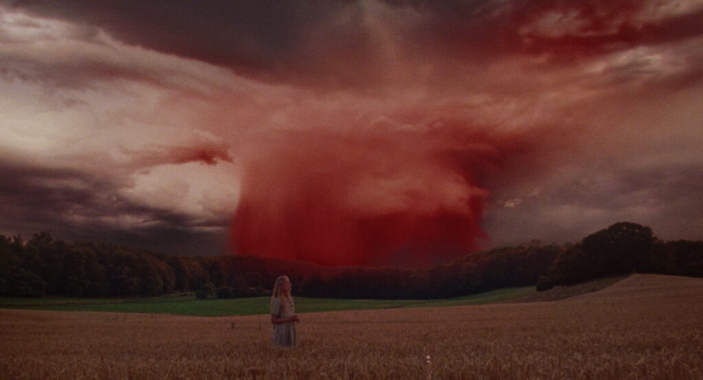 Young woman standing in a wheat field beneath an enormous red storm cloud spreading across the sky.