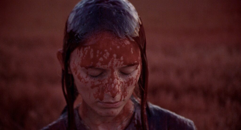Close-up of a young woman with blood covering her face against a deep red background.